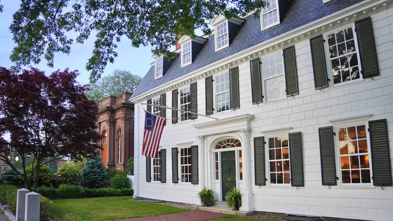 A white colonial house with dark shutters at dusk, lights on inside. Reference photograph.