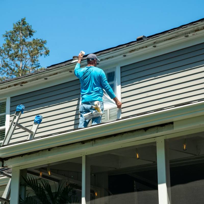 A painter on a ladder masking a window on grey clapboard siding. Reference photograph.
